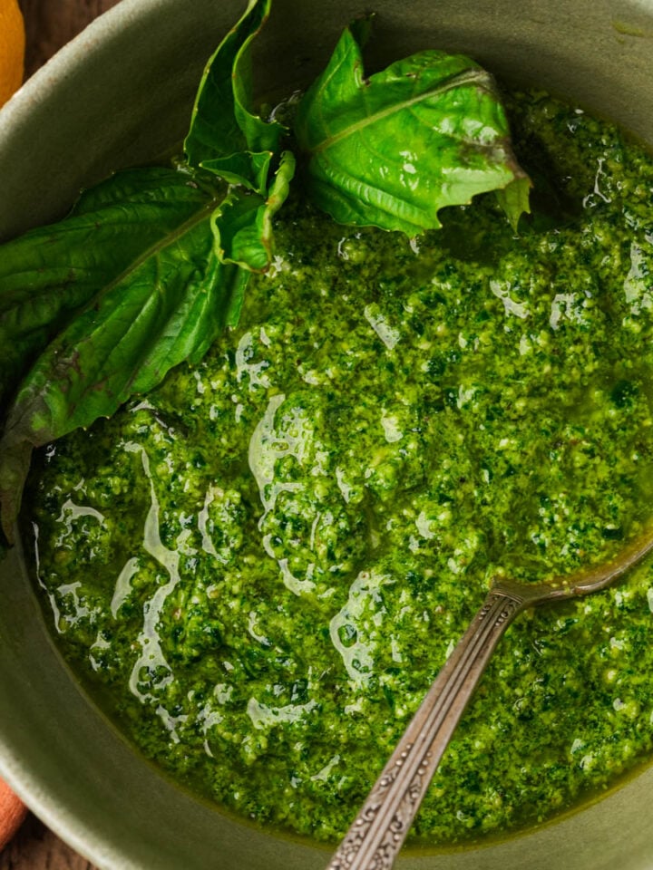 A bowl of spinach pesto garnished with fresh basil leaves, with a spoon in the bowl. Part of a lemon and a small dish of coarse salt are visible beside the bowl.