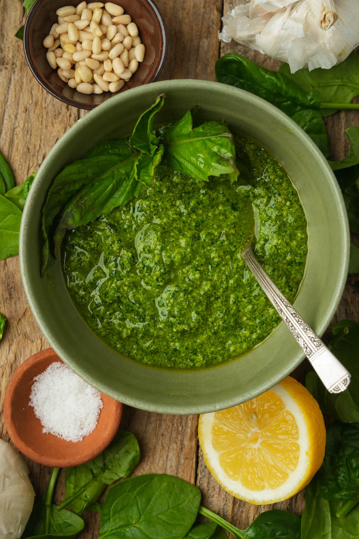 A green bowl of spinach pesto with a spoon, surrounded by fresh basil leaves, pine nuts, garlic, coarse salt, spinach, and a halved lemon on a rustic wooden surface.