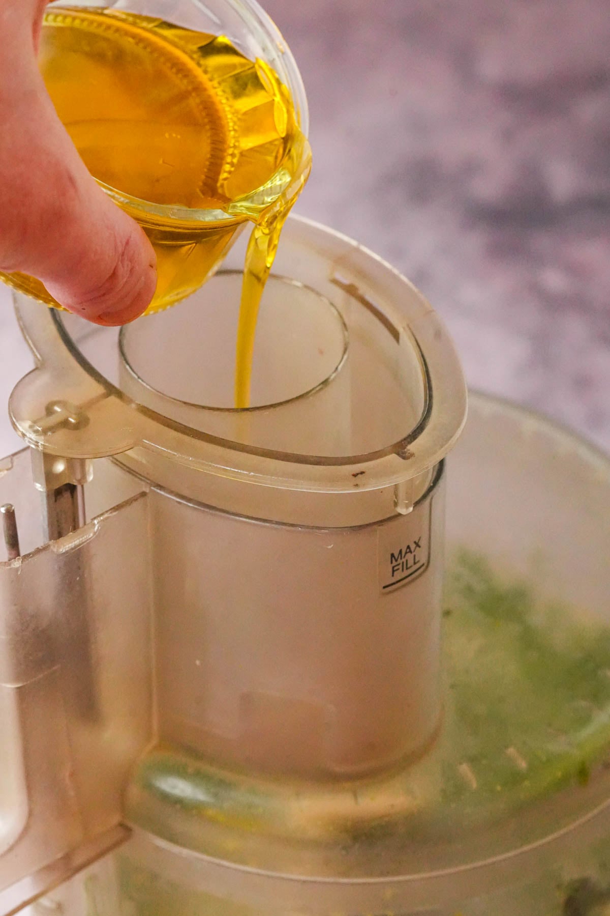 A hand pours olive oil from a small glass bowl into the top opening of a food processor, which contains blending spinach pesto.