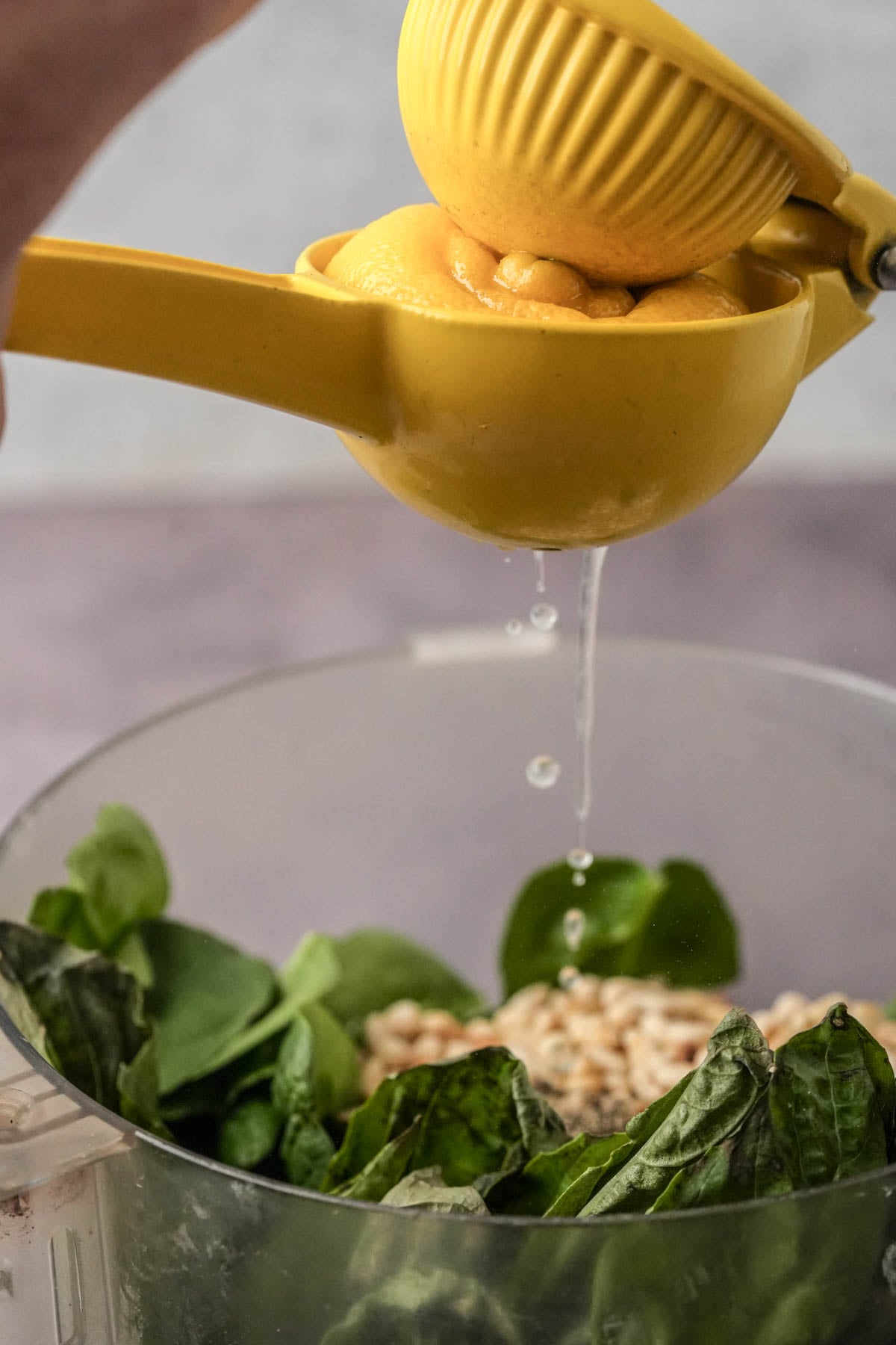 A hand squeezes a yellow lemon squeezer over a food processor filled with ingredients for a spinach pesto recipe.