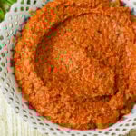 A bowl filled with a thick, sun dried tomato pesto, speckled with green herbs, served in a decorative white dish with a lace-like rim.