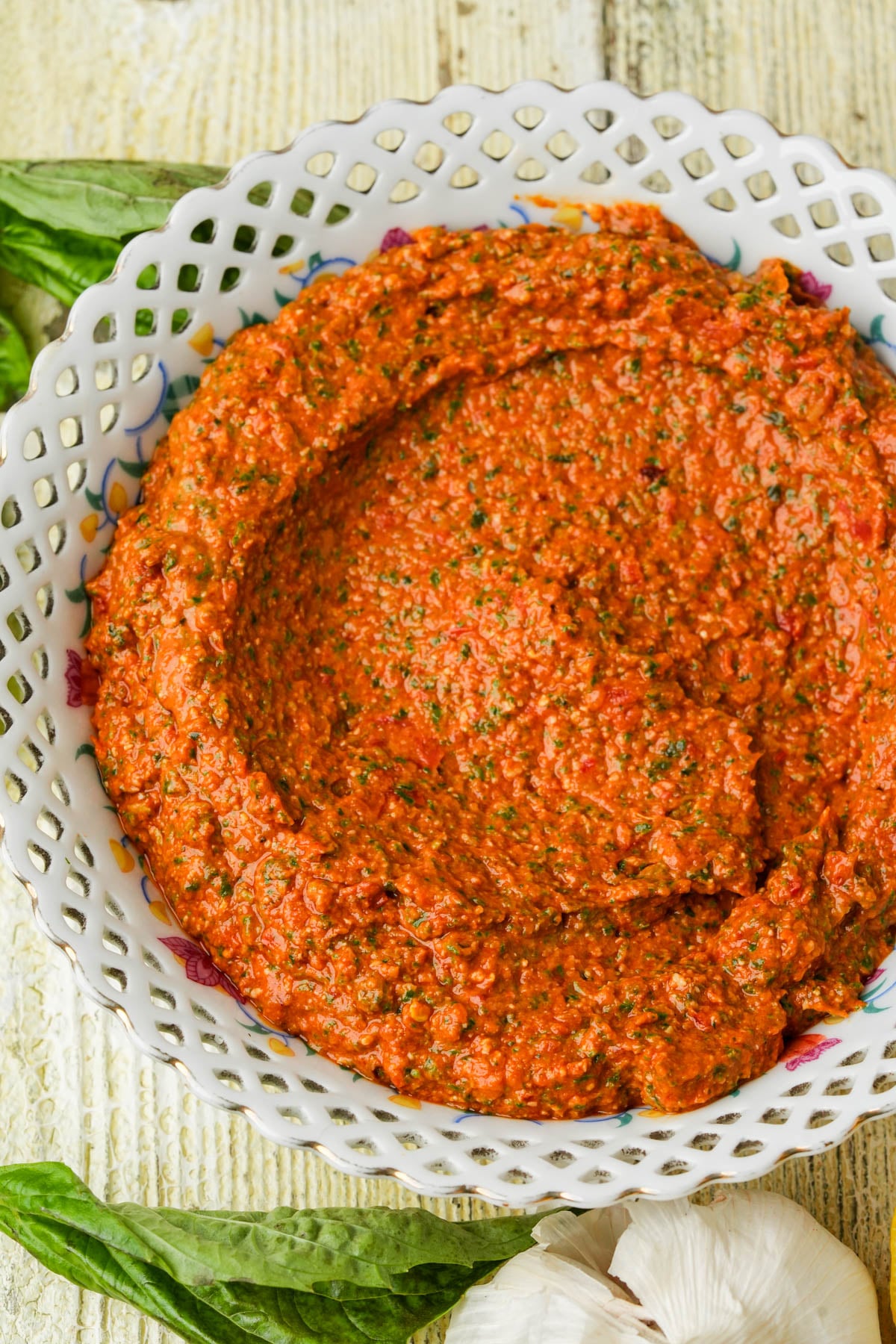 A decorative white bowl filled with sun dried tomato pesto sits on a wooden surface, garnished with green herbs. Fresh basil leaves and garlic bulbs are nearby.