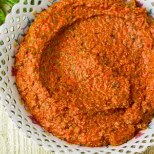 A bowl filled with a thick, sun dried tomato pesto, speckled with green herbs, served in a decorative white dish with a lace-like rim.