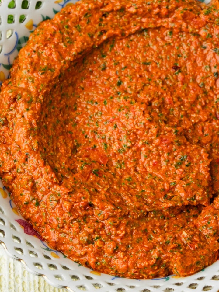 A bowl filled with a thick, sun dried tomato pesto, speckled with green herbs, served in a decorative white dish with a lace-like rim.