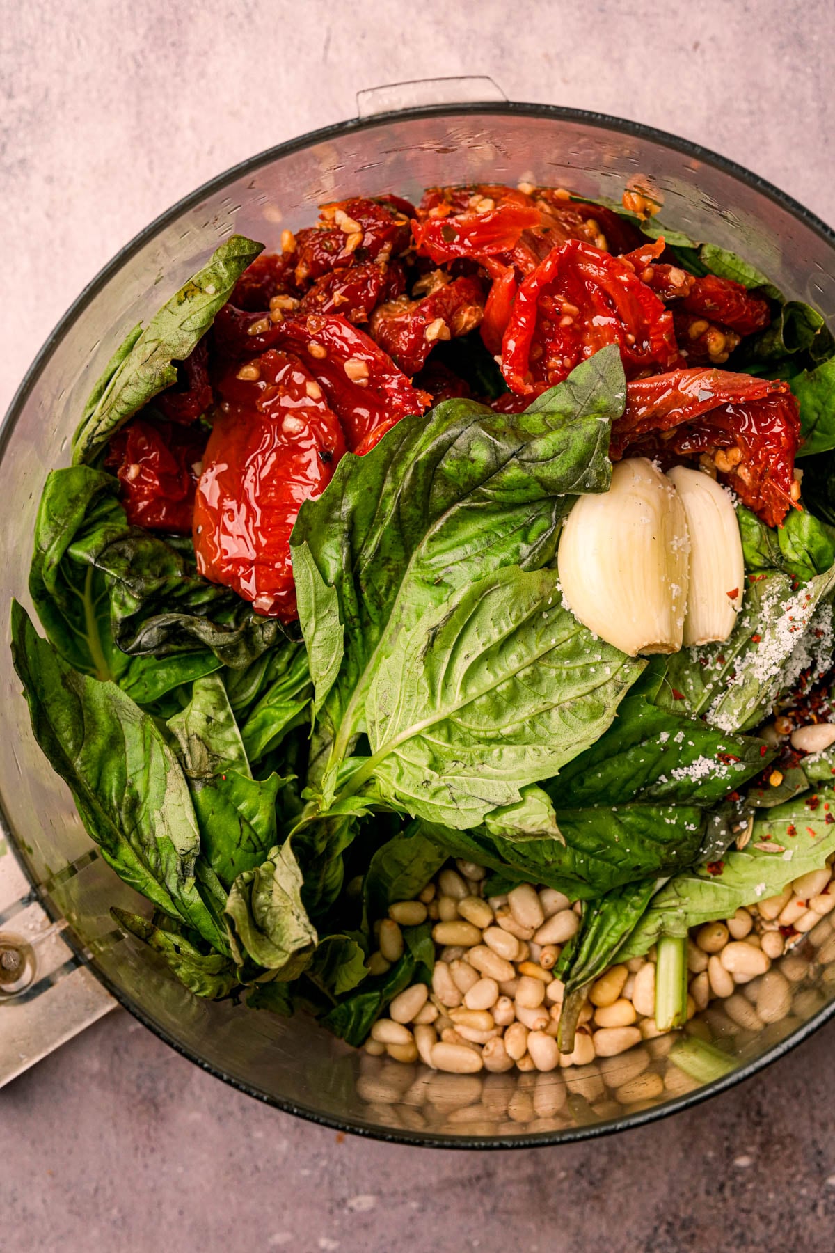 A food processor bowl filled with fresh basil leaves, sun-dried tomatoes, garlic cloves, pine nuts, and a sprinkle of salt, ready to be blended to become some great sun dried tomato pesto.