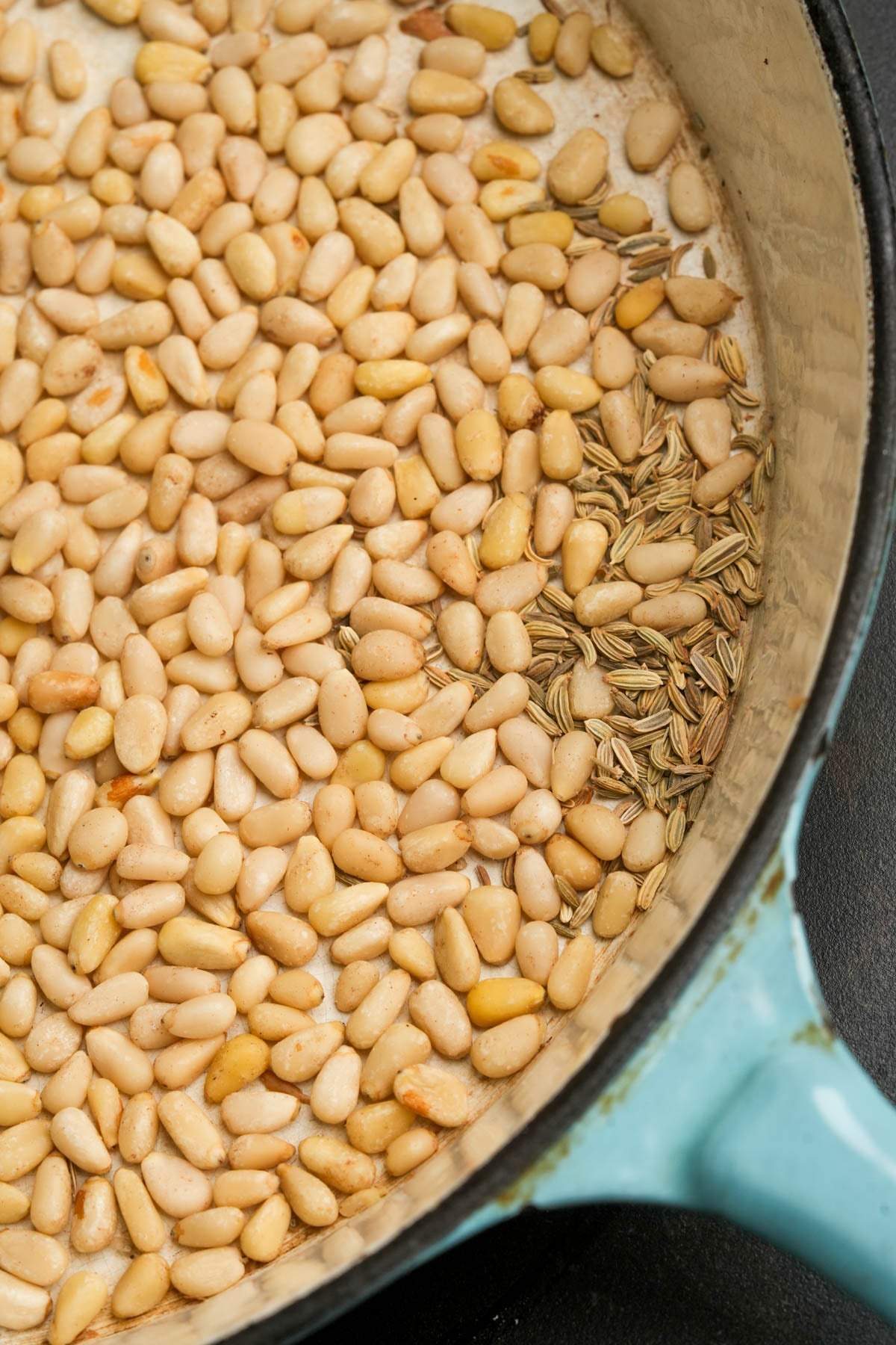A close-up of a blue pot with pine nuts and fennel seeds toasting inside, seen from above on a dark surface.