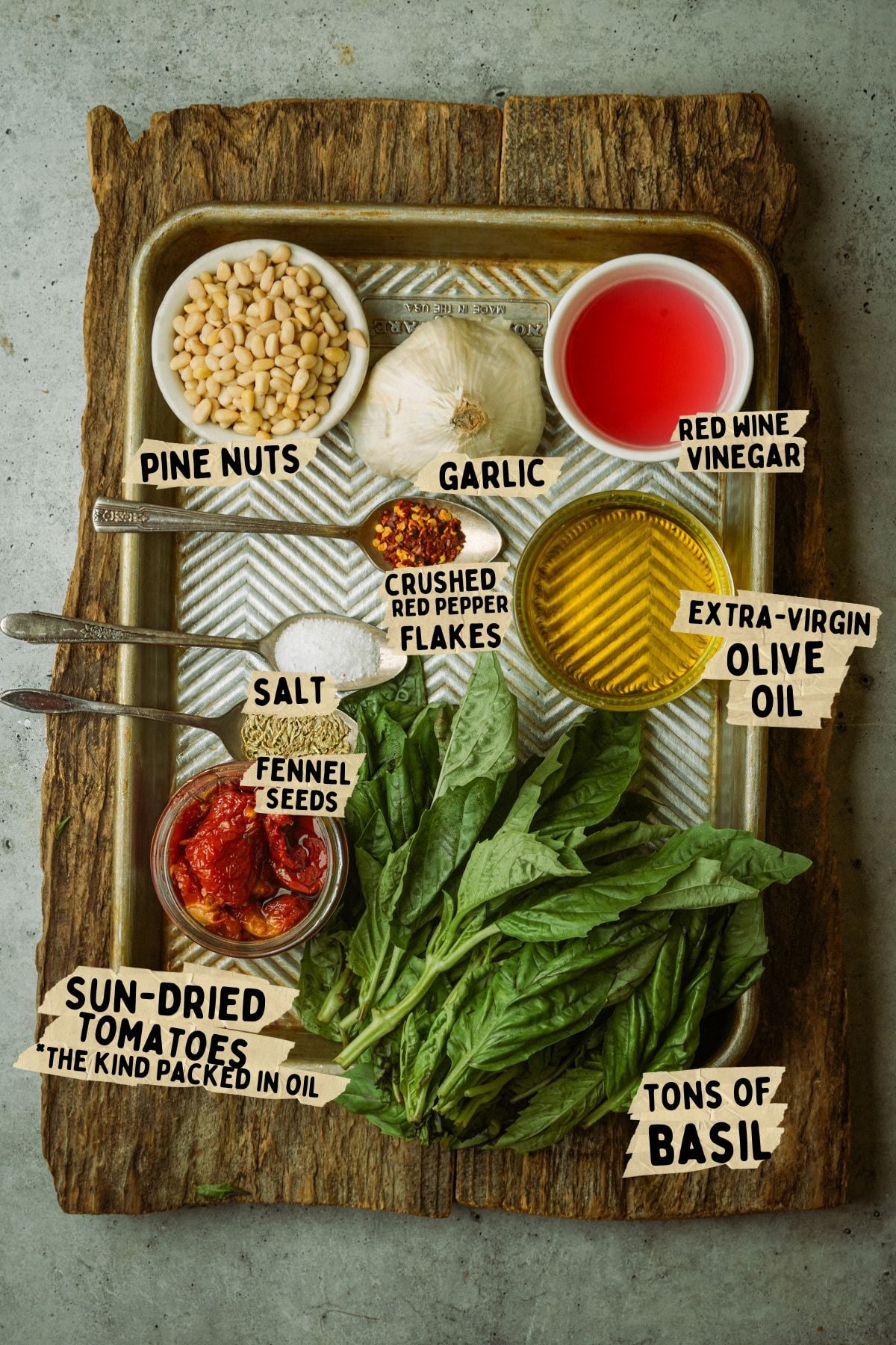A tray with labeled ingredients for a sun dried tomato pesto recipe: pine nuts, garlic, red wine vinegar, extra-virgin olive oil, salt, crushed red pepper flakes, fennel seeds, sun-dried tomatoes in oil, and lots of fresh basil.