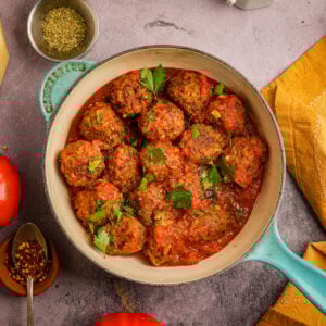 Tofu meatballs in tomato sauce garnished with fresh parsley, served in a blue Dutch oven on a stone surface, surrounded by seasonings, a tomato, and a yellow napkin.