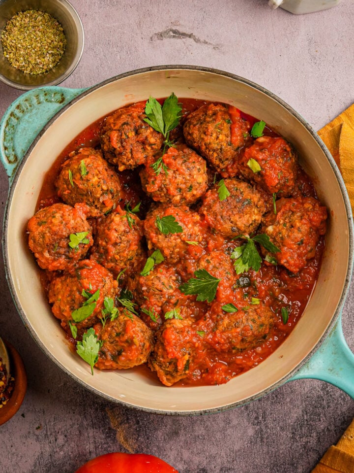 Tofu meatballs in tomato sauce garnished with fresh parsley, served in a blue Dutch oven on a stone surface, surrounded by seasonings, a tomato, and a yellow napkin.