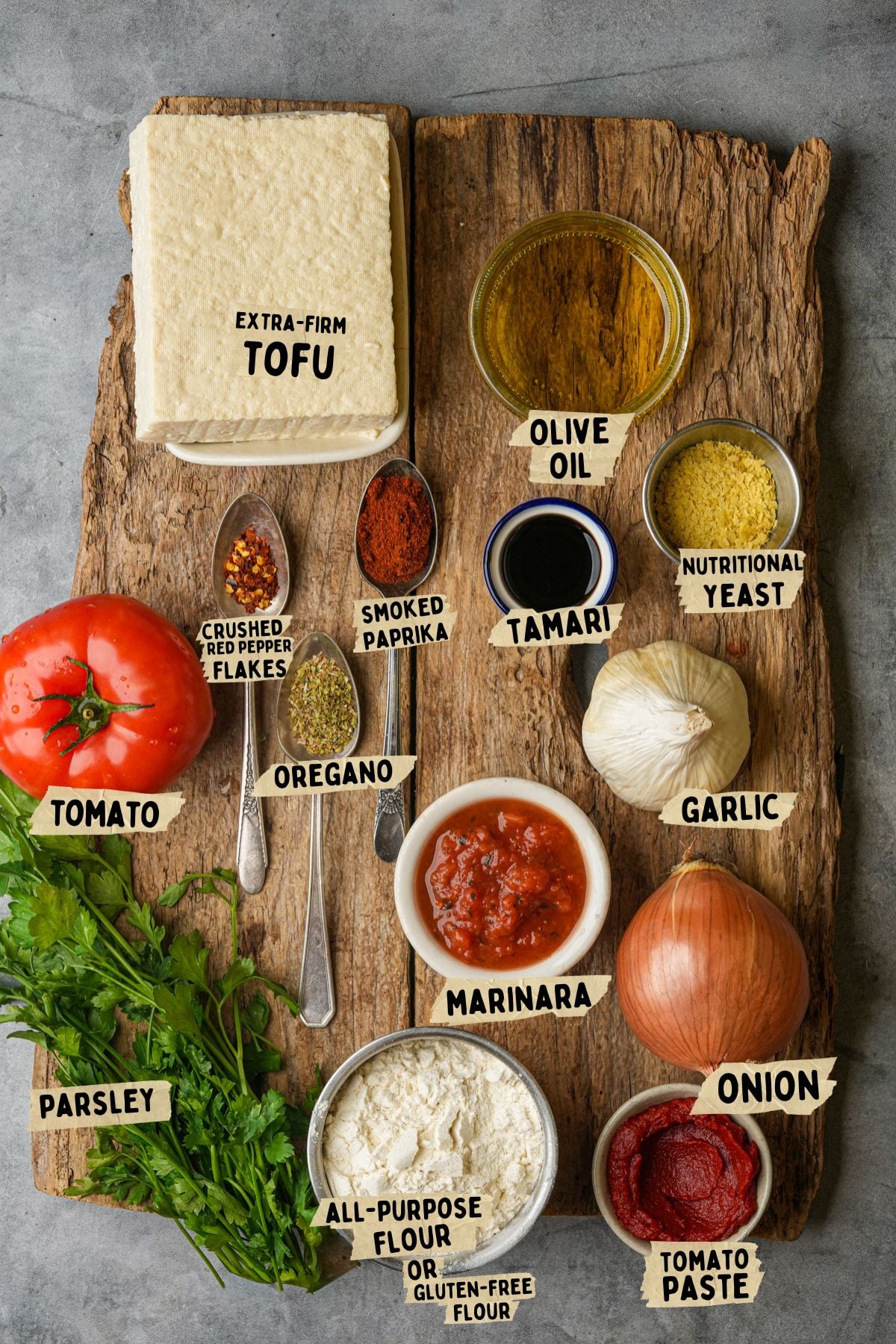 A wooden board displays labeled ingredients for a tofu meatballs recipe: extra-firm tofu, olive oil, tamari, nutritional yeast, garlic, onion, tomato paste, marinara sauce, all-purpose flour, parsley, a tomato, oregano, crushed red pepper flakes, and smoked paprika.