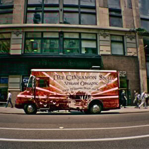 A red and white food truck labeled The Cinnamon Snail Vegan Organic is parked on a city street in front of a tall building, with several pedestrians walking by on the sidewalk.