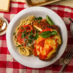A white plate on a red-checkered tablecloth holds spaghetti with marinara sauce and grated cheese, next to a piece of vegan chicken parmesan topped with melted cheese and tomato sauce, garnished with fresh herbs.