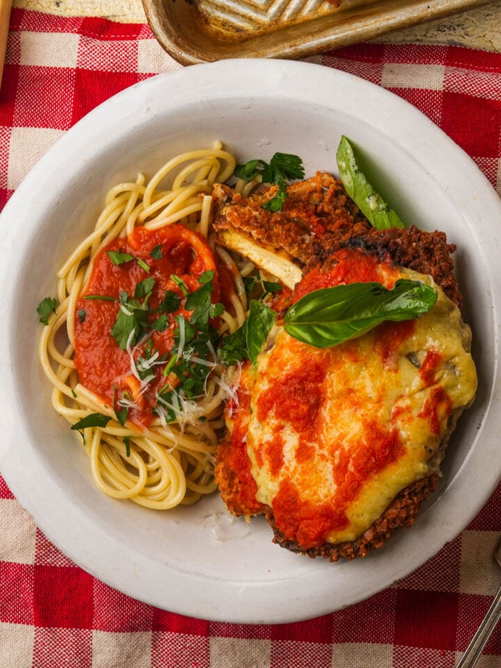A white plate on a red-checkered tablecloth holds spaghetti with marinara sauce and grated cheese, next to a piece of vegan chicken parmesan topped with melted cheese and tomato sauce, garnished with fresh herbs.