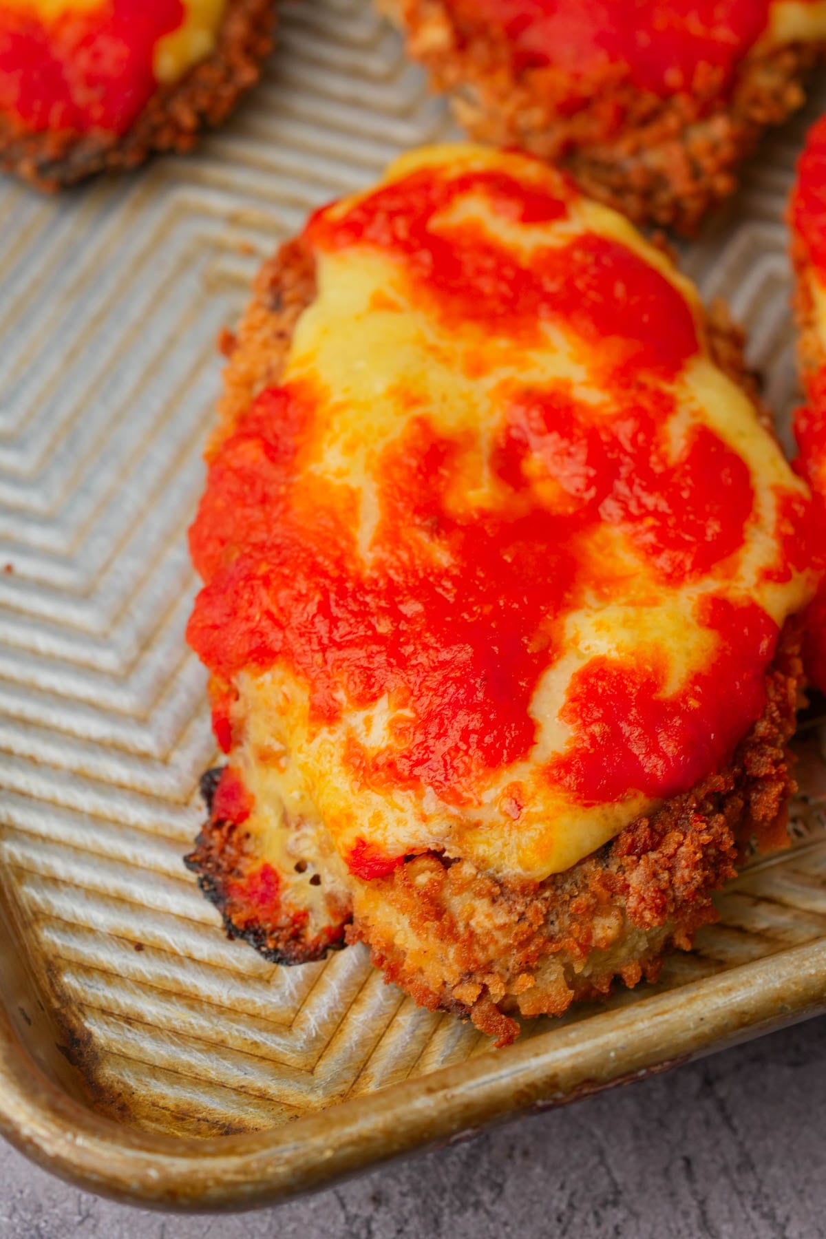 A close-up of a breaded tofu chicken cutlet topped with melted cheese and tomato sauce on a metal baking sheet.