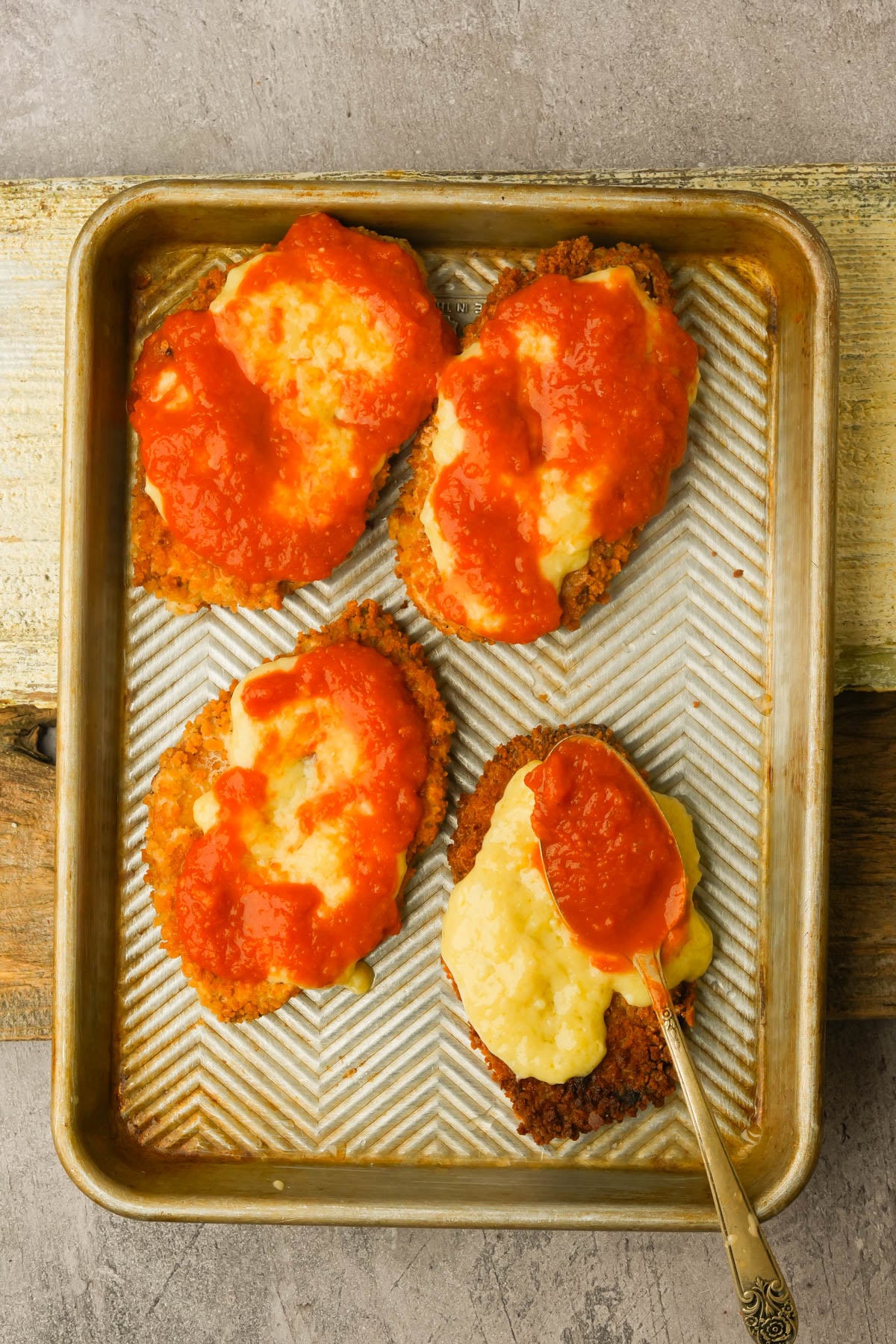 A baking tray with four breaded tofu cutlets topped with melted cheese and tomato sauce, one with a spoon resting on it. The tray sits on a rustic wooden surface.
