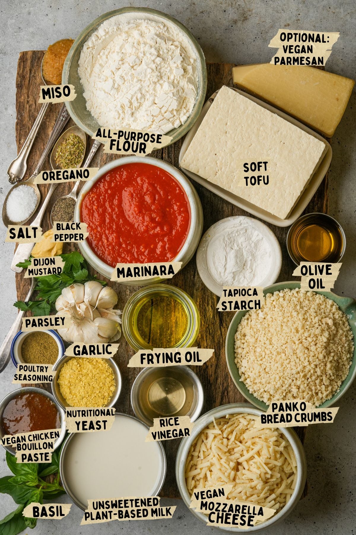 An assortment of labeled ingredients on a table for a vegan chicken parmesan recipe including all-purpose flour, tofu, marinara sauce, bread crumbs, vegan cheeses, spices, oil, garlic, plant milk, and nutritional yeast.
