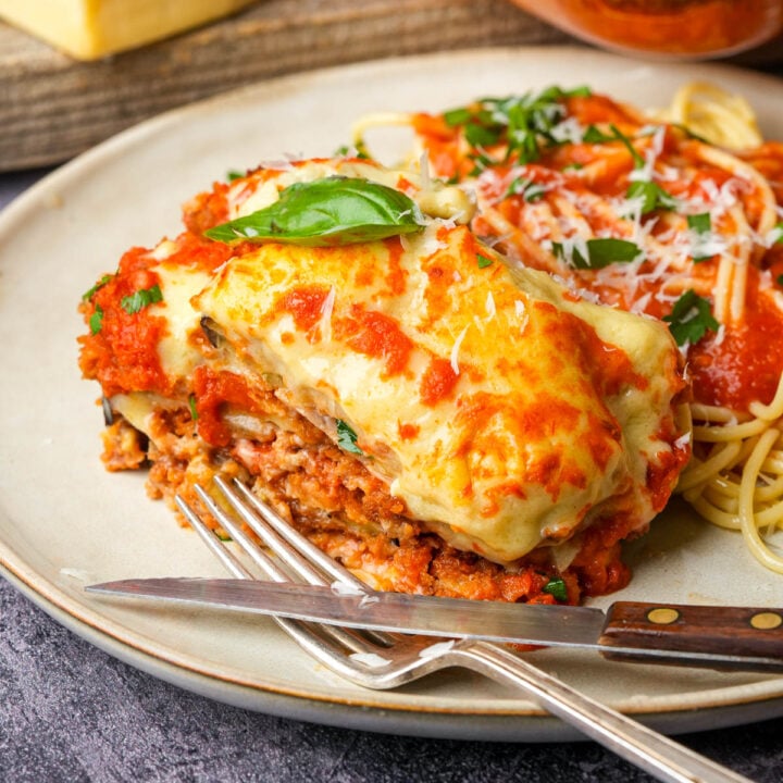 A close-up of a plate with cheesy vegan eggplant parm topped with fresh basil and marinara sauce, next to spaghetti garnished with parsley and grated cheese; a fork and knife rest beside the food.
