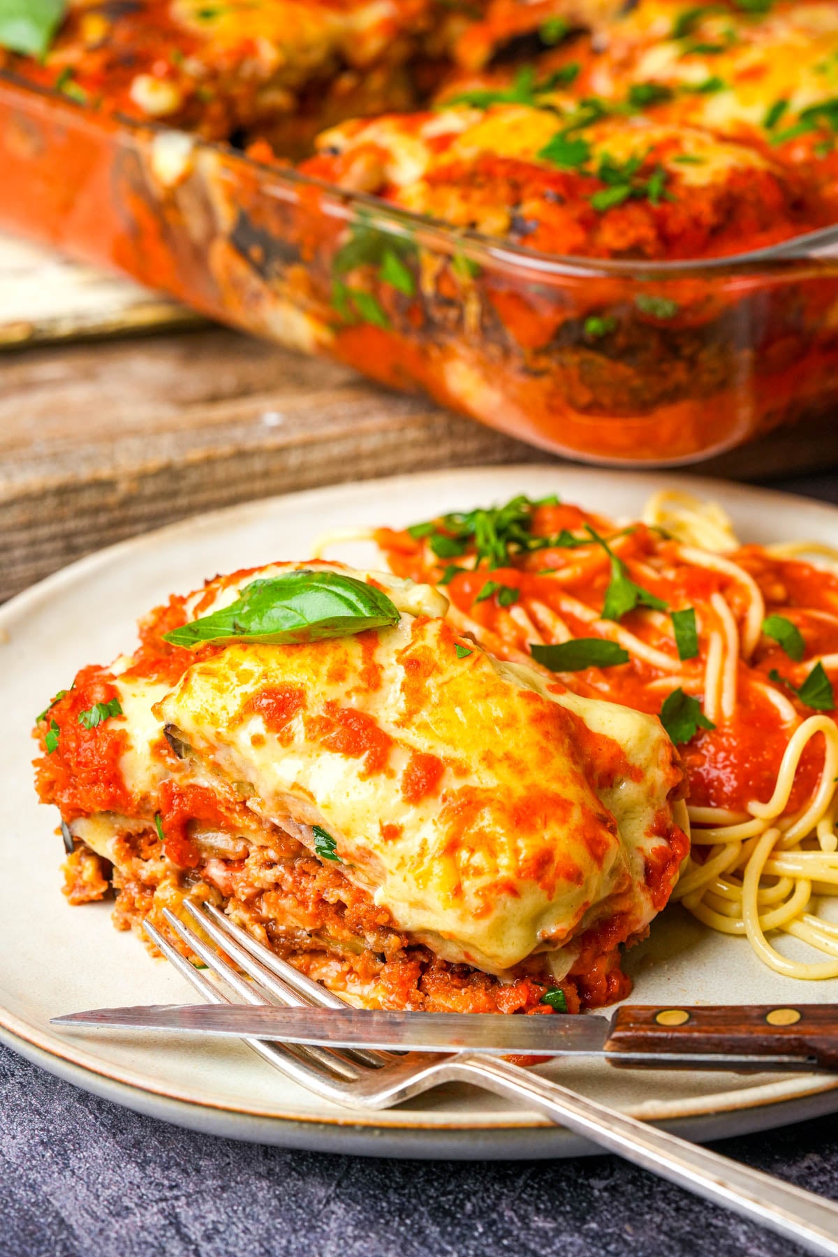 A plate with a serving of cheesy baked vegan eggplant parm next to spaghetti topped with tomato sauce and fresh basil, with a fork and knife beside it. A baking dish with more parm is in the background.