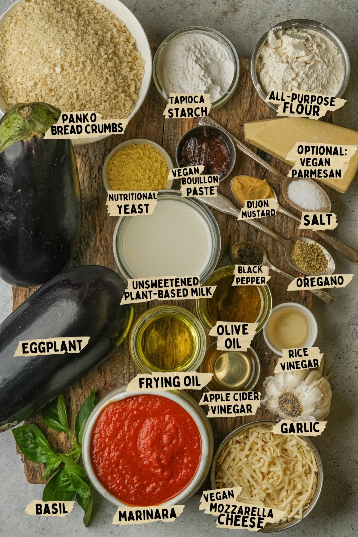 A flat lay of ingredients for a vegan eggplant parm recipe including eggplant, marinara, garlic, vegan mozzarella, panko crumbs, flour, starch, spices, nutritional yeast, vegan bouillon, olive oil, frying oil, oregano, and basil.