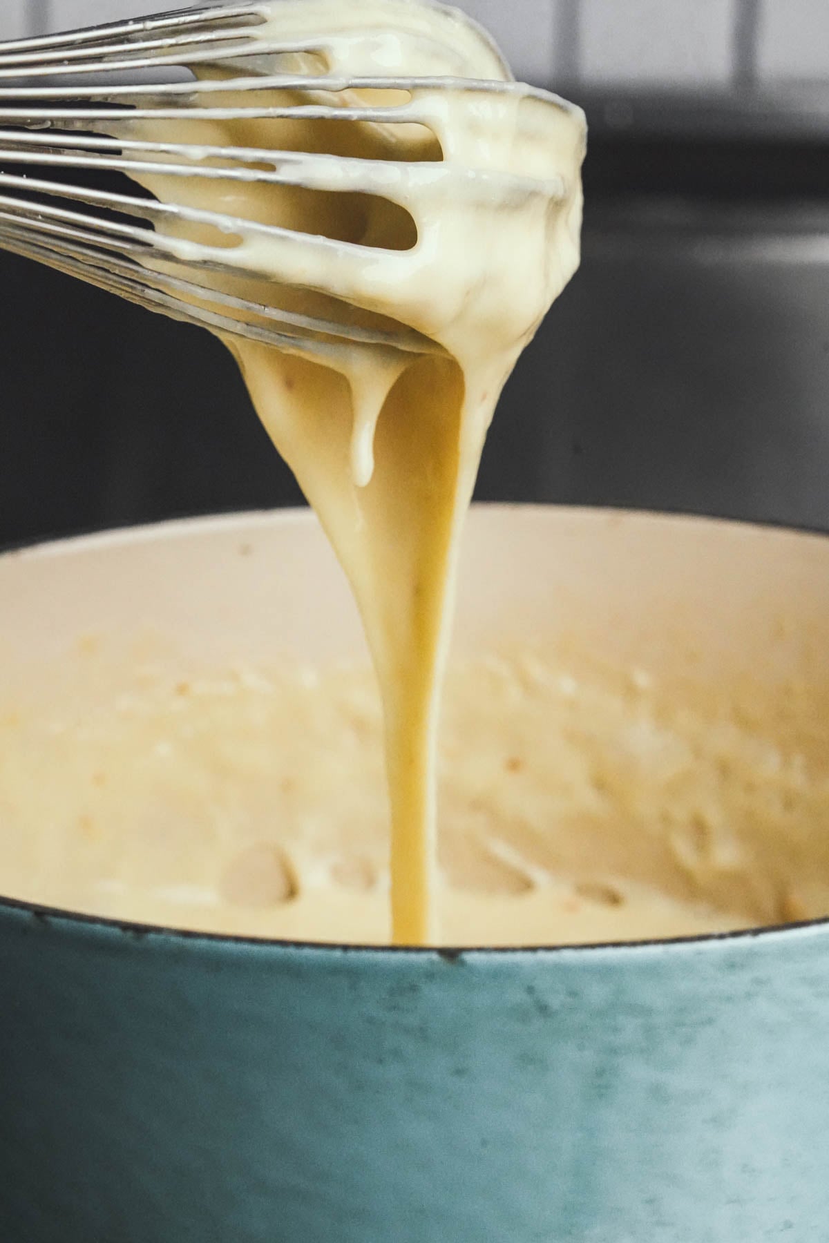 A metal whisk lifting up thick, creamy vegan cheese sauce from a blue pot, with the sauce dripping smoothly back into the pot.