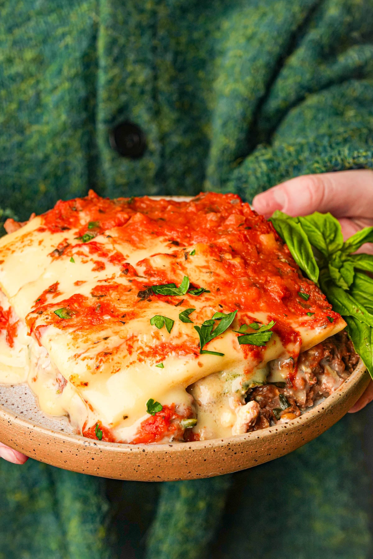 A person in a green sweater holds a plate of cheesy baked vegan lasagna topped with tomato sauce and garnished with fresh parsley and basil.