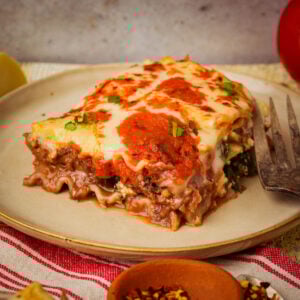 A slice of vegan lasagna with layers of pasta, lentil meat, melted cheese, and herbs sits on a beige plate beside a fork, with a red and white striped napkin underneath.