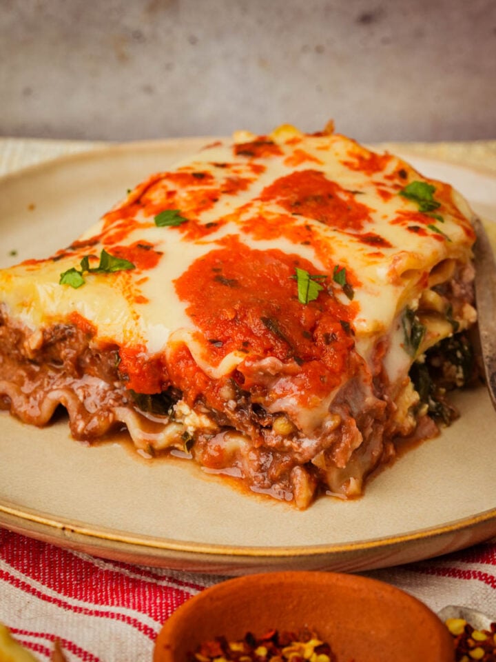 A slice of vegan lasagna with layers of pasta, lentil meat, melted cheese, and herbs sits on a beige plate beside a fork, with a red and white striped napkin underneath.