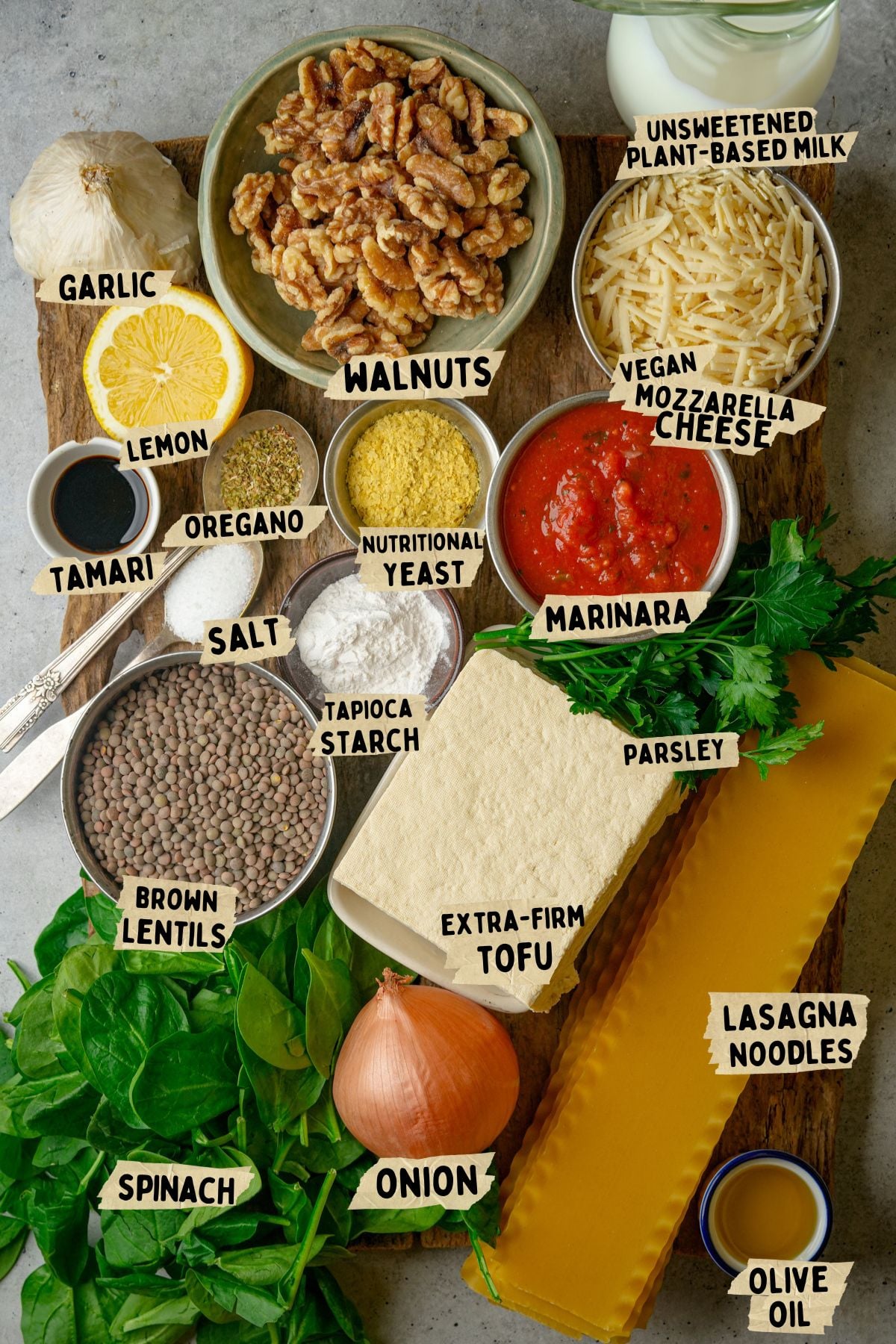 A variety of vegan lasagna recipe ingredients on a wooden board, including walnuts, tofu, spinach, lentils, onion, parsley, vegan mozzarella cheese, marinara sauce, lasagna noodles, and various seasonings.