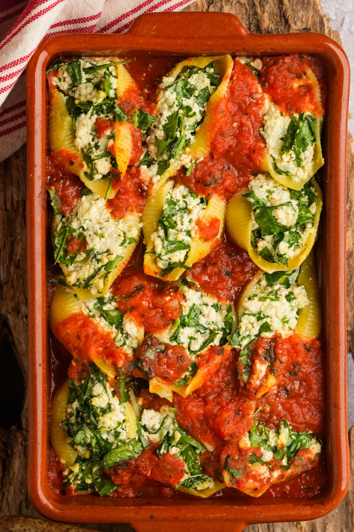 A brown baking dish filled with jumbo pasta shells stuffed with vegan ricotta and spinach, topped with tomato sauce. The shells are arranged closely together, and some spinach and cheese filling is visible. A striped towel is in the corner.