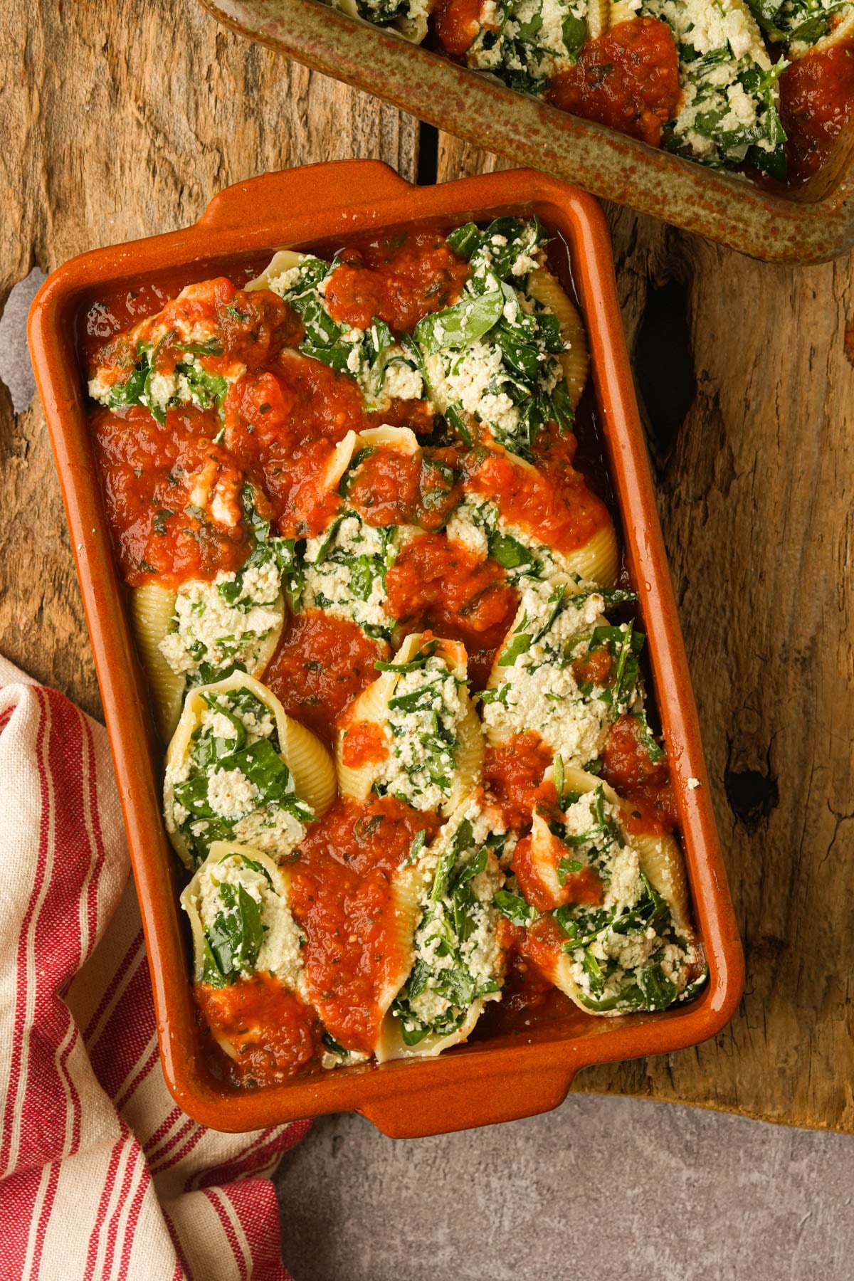 A rectangular baking dish filled with jumbo pasta shells stuffed with vegan ricotta and spinach, topped with tomato sauce, sits on a rustic wooden surface next to a red and white striped cloth.