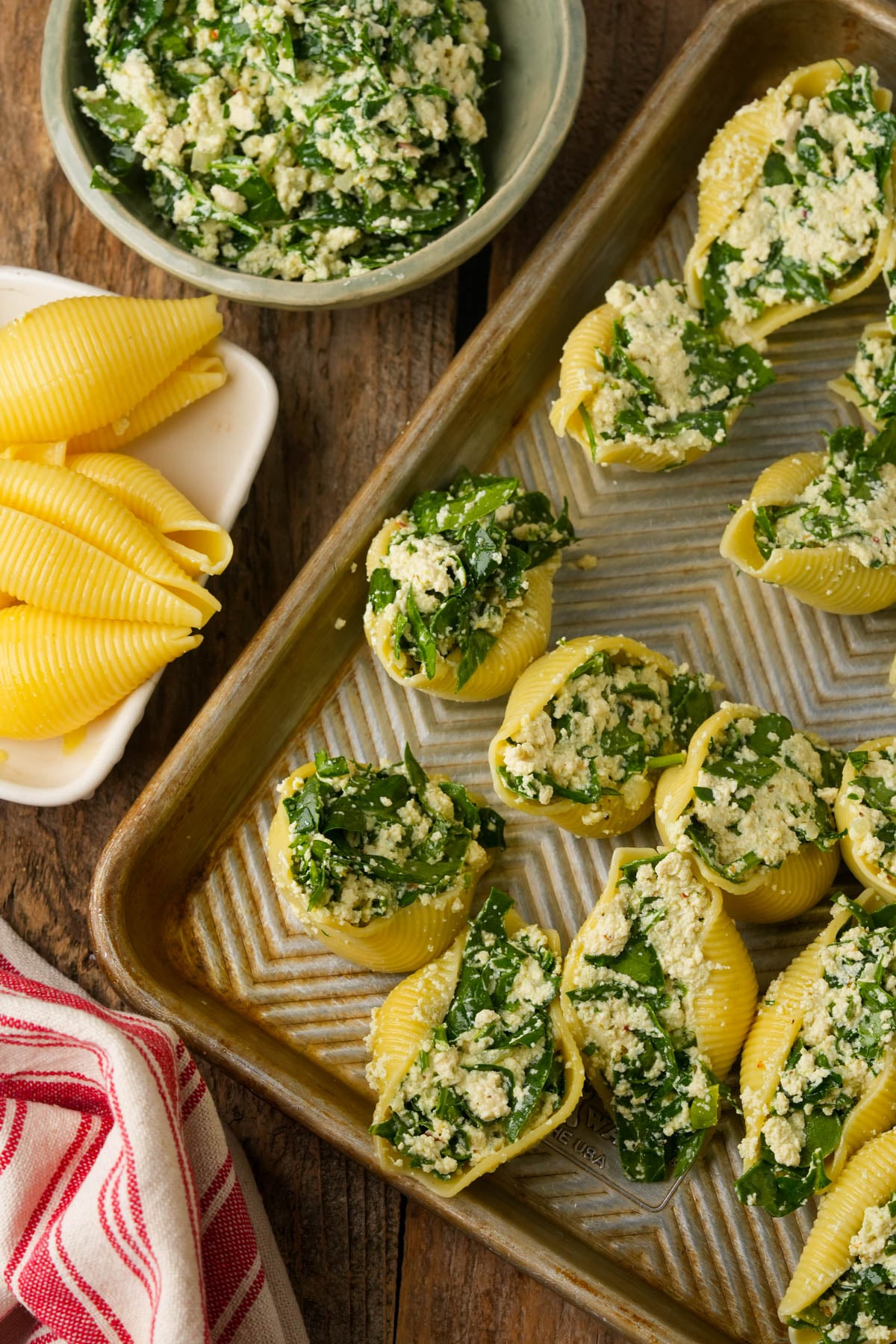A baking tray with jumbo pasta shells stuffed with a spinach and cheese mixture. Unfilled pasta shells sit nearby, along with a bowl of extra filling and a red-striped kitchen towel on a rustic wooden table.