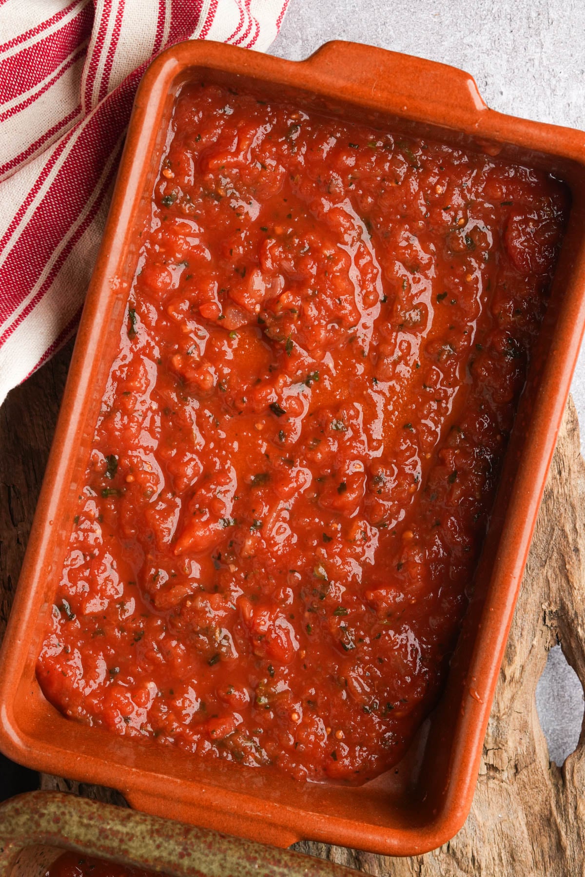 A rectangular ceramic dish filled with chunky tomato sauce, garnished with herbs, sits on a wooden surface next to a red and white striped cloth.