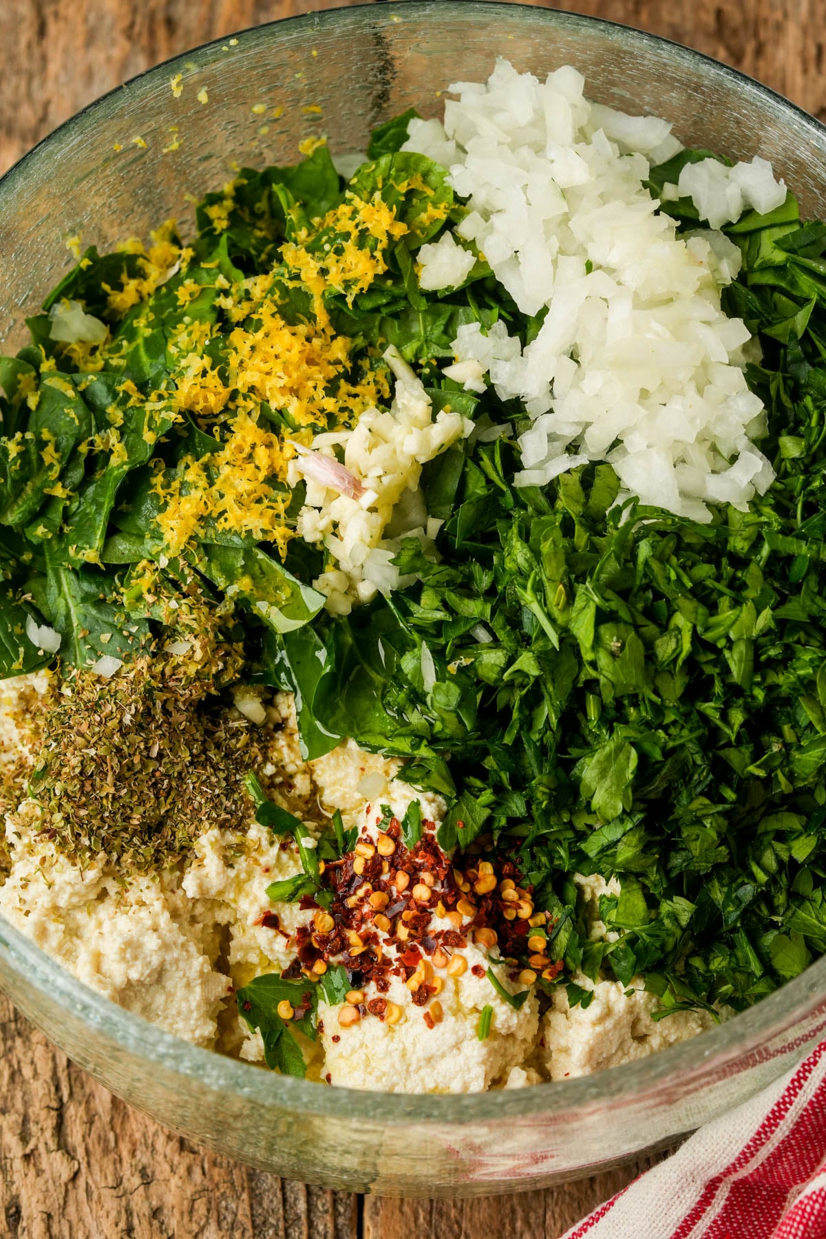 A glass bowl filled with began ricotta cheese, chopped onions, parsley, spinach, lemon zest, dried oregano, and red pepper flakes on a wooden surface. A red and white towel is partially visible in the corner.