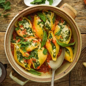 A rustic bowl filled with vegan stuffed shells with ricotta, spinach, and herbs, topped with tomato sauce and fresh basil leaves, set on a wooden table with a serving spoon.