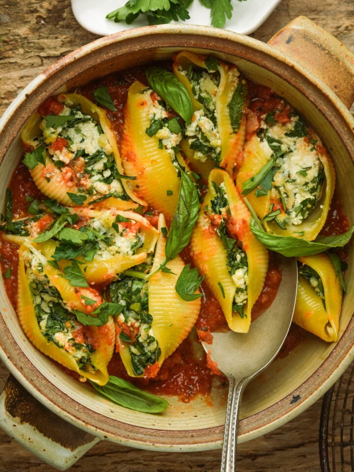 A rustic bowl filled with vegan stuffed shells with ricotta, spinach, and herbs, topped with tomato sauce and fresh basil leaves, set on a wooden table with a serving spoon.