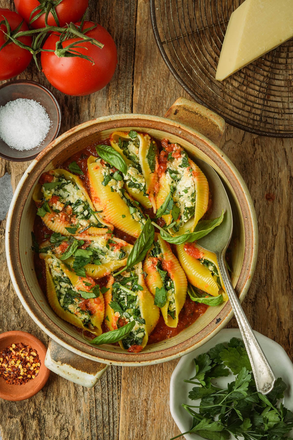 A bowl of vegan stuffed shells filled with ricotta, spinach, and herbs, topped with tomato sauce and fresh basil. Surrounding the bowl are tomatoes, a wedge of cheese, salt, red pepper flakes, and fresh parsley.