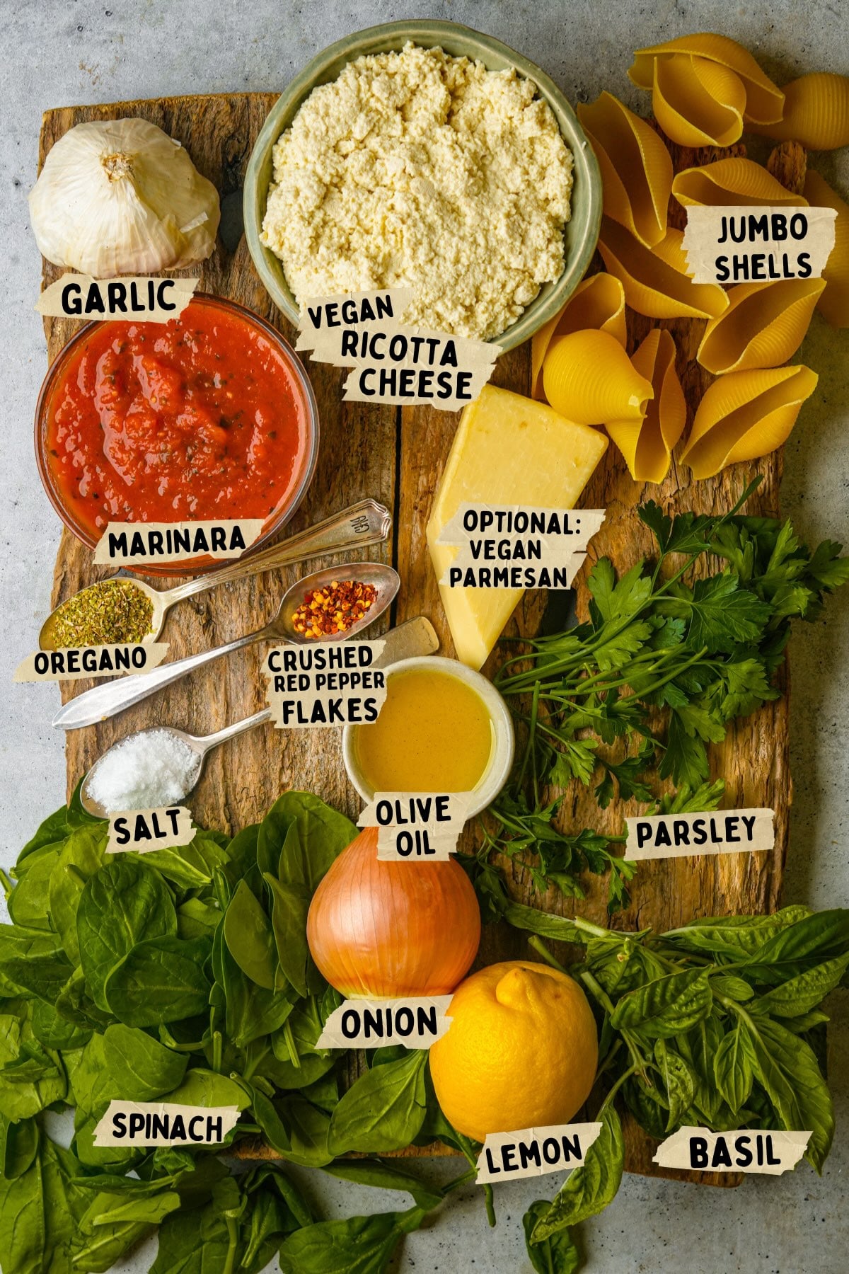 A wooden board with labeled ingredients for a vegan stuffed shells recipe: garlic, vegan ricotta cheese, jumbo shells, marinara, oregano, salt, crushed red pepper flakes, vegan parmesan, olive oil, parsley, onion, lemon, spinach, and basil.