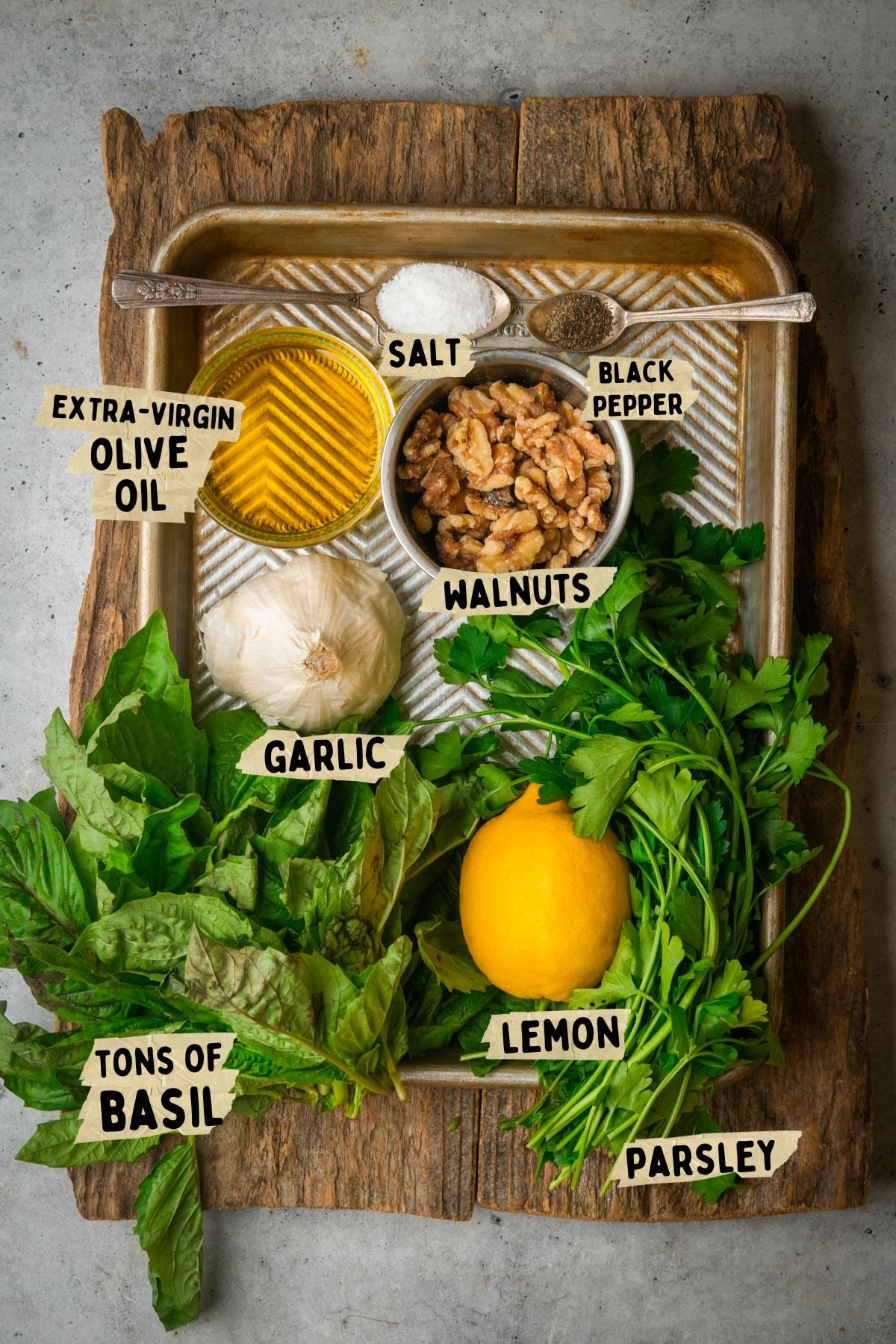 A tray holds labeled ingredients for a walnut pesto recipe: extra-virgin olive oil, salt, black pepper, walnuts, garlic, lots of fresh basil, a lemon, and parsley, all arranged on a wooden board.