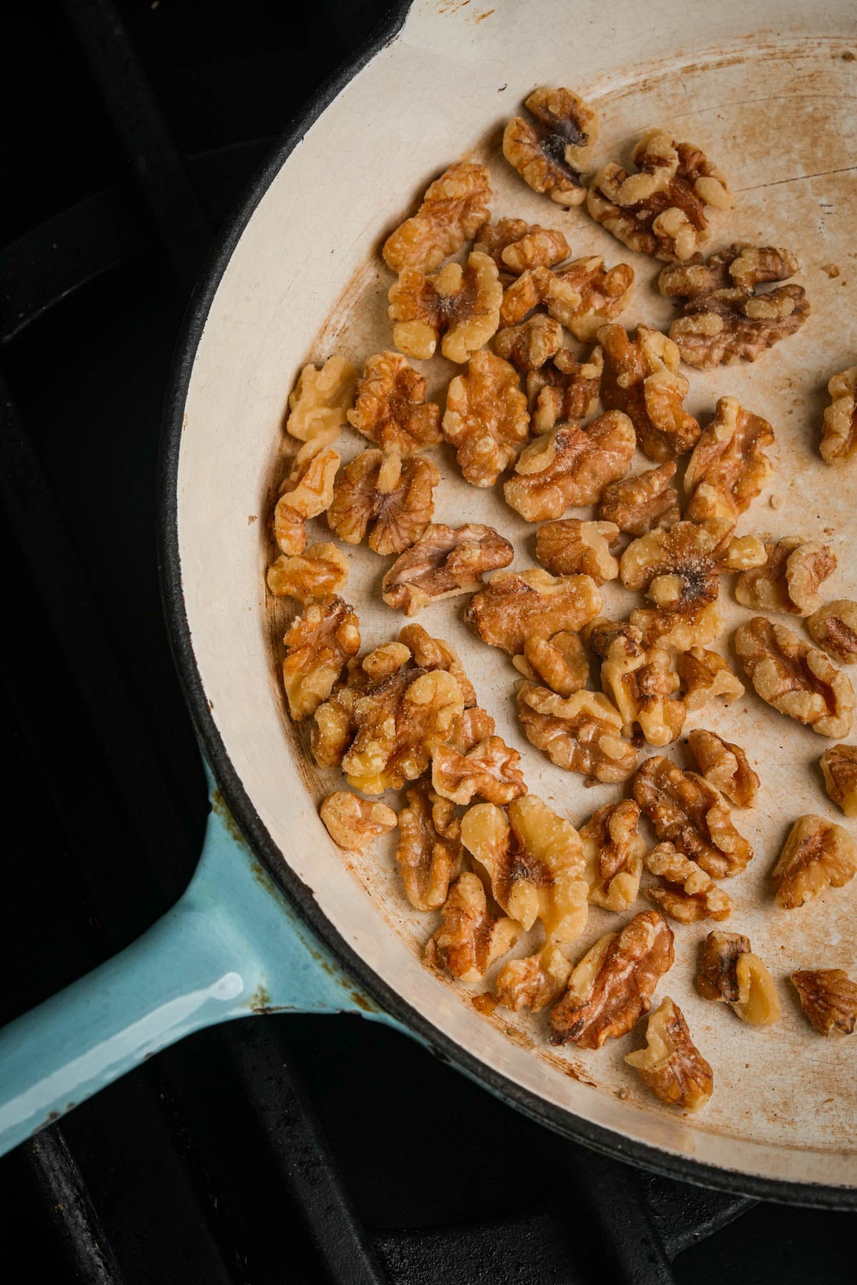 A close-up of walnut halves being toasted in a light-colored pan with a blue handle on a stovetop.