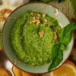 A bowl of walnut pesto sauce garnished with chopped walnuts and fresh basil leaves, next to a lemon wedge and a clove of garlic, on a rustic wooden surface.