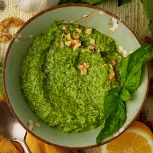 A bowl of walnut pesto sauce garnished with chopped walnuts and fresh basil leaves, next to a lemon wedge and a clove of garlic, on a rustic wooden surface.