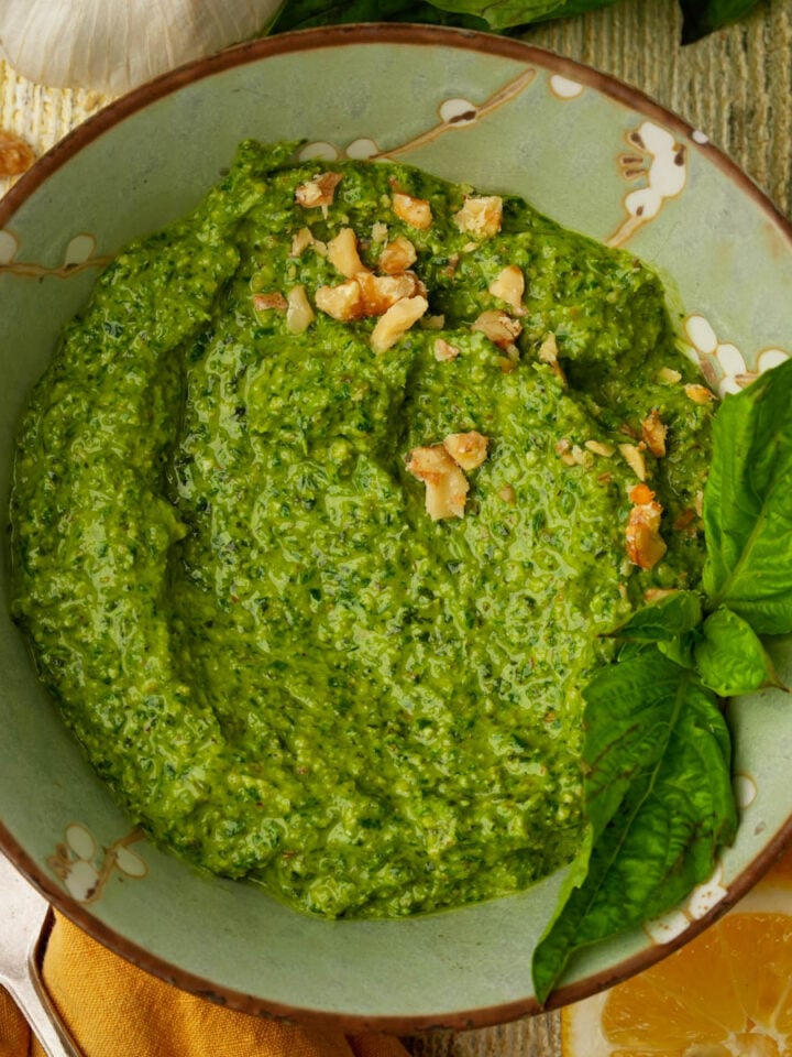 A bowl of walnut pesto sauce garnished with chopped walnuts and fresh basil leaves, next to a lemon wedge and a clove of garlic, on a rustic wooden surface.