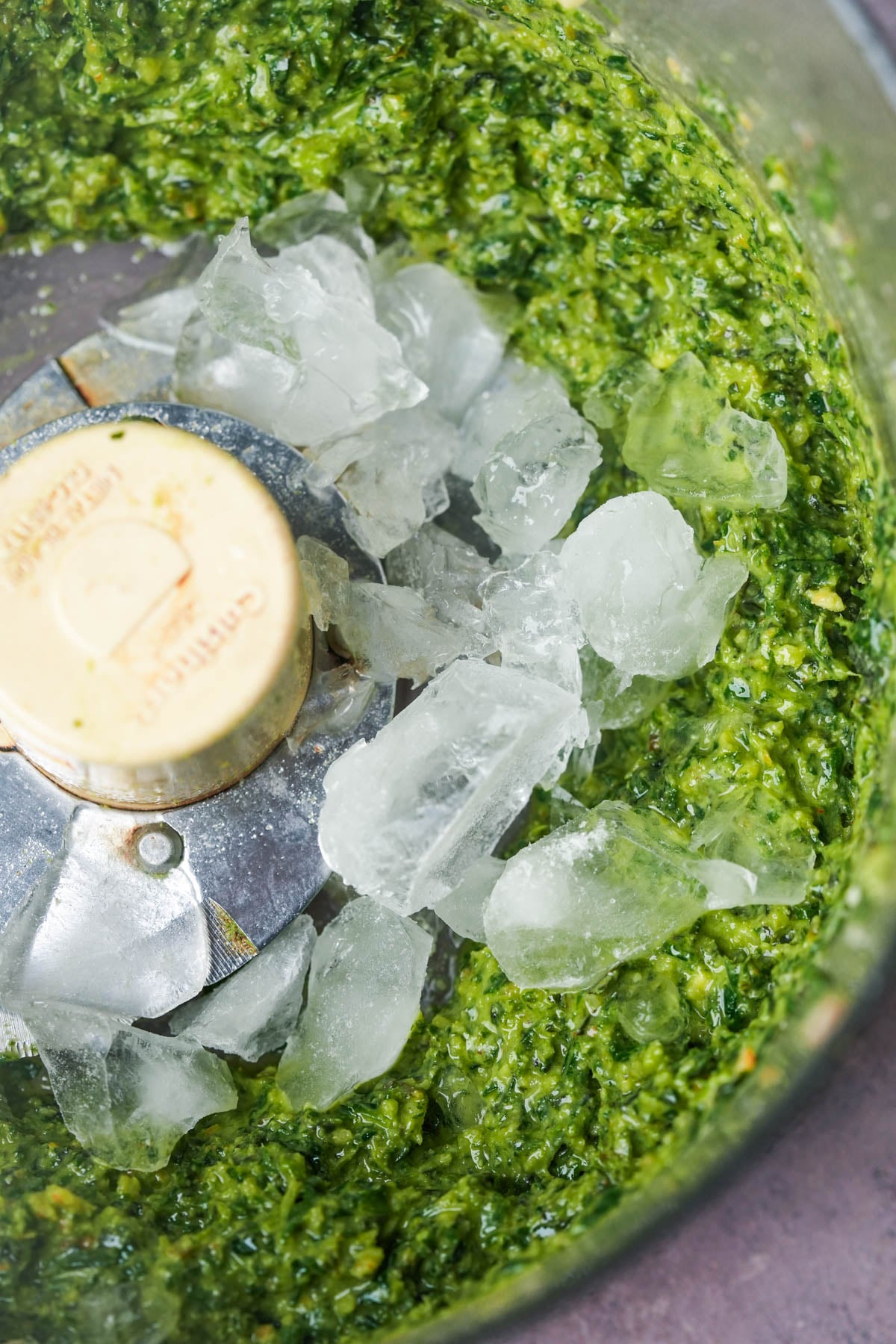 A food processor bowl containing bright green walnut pesto sauce with several ice cubes on top of the mixture, ready to be blended further.