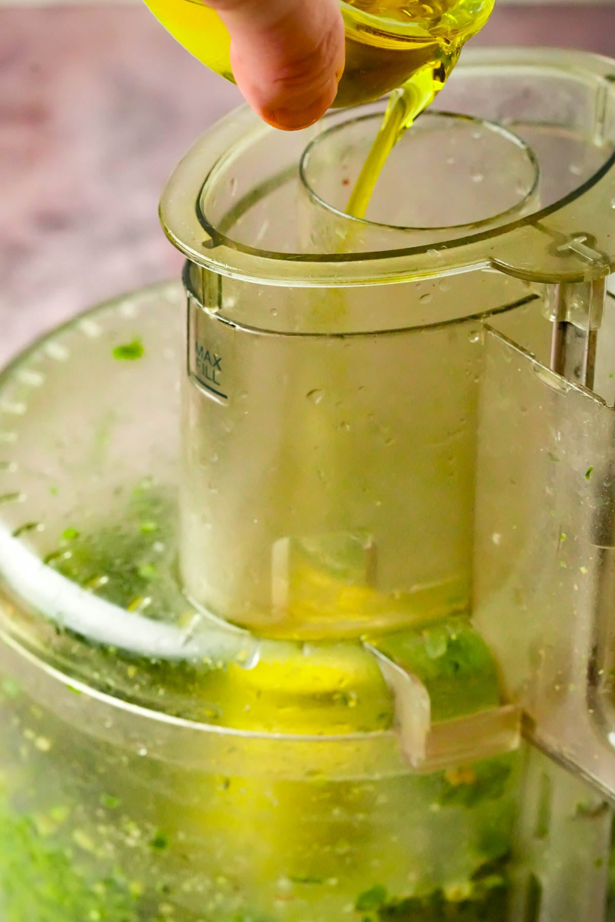A food processor blending walnut pesto while someone pours olive oil from a small container through the top opening.