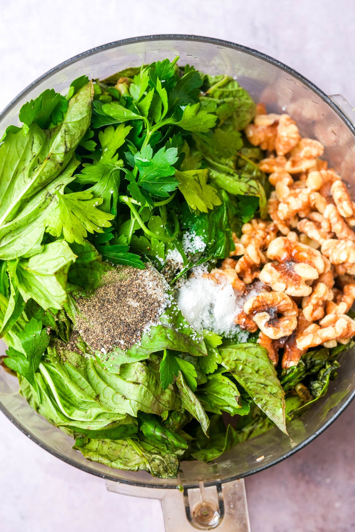 A food processor bowl filled with ingredients for a walnut pesto recipe including fresh basil leaves, parsley, walnuts, salt, and pepper, ready to be blended.