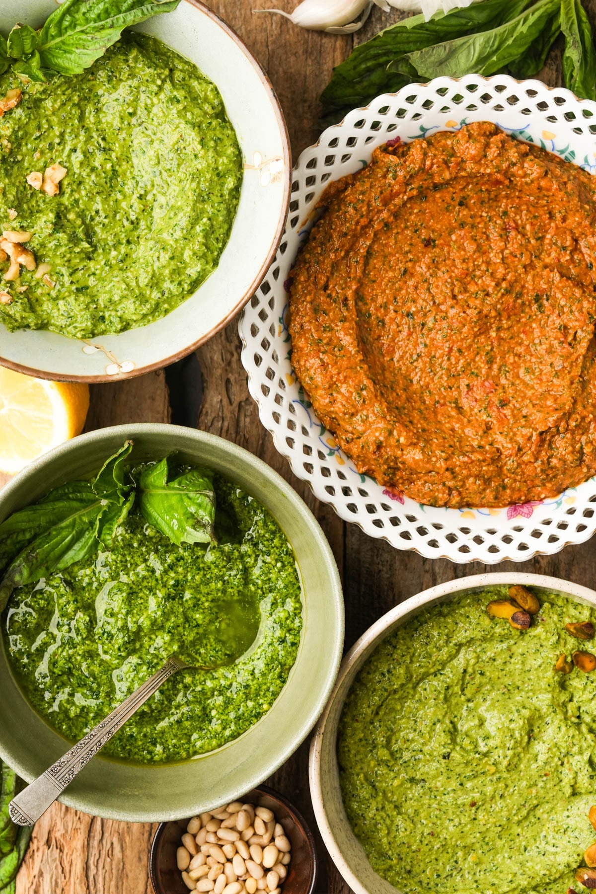 Four bowls of pesto sauce: walnut pesto, pistachio pesto, sun dried tomato pesto, and spinach pesto on a wooden table.