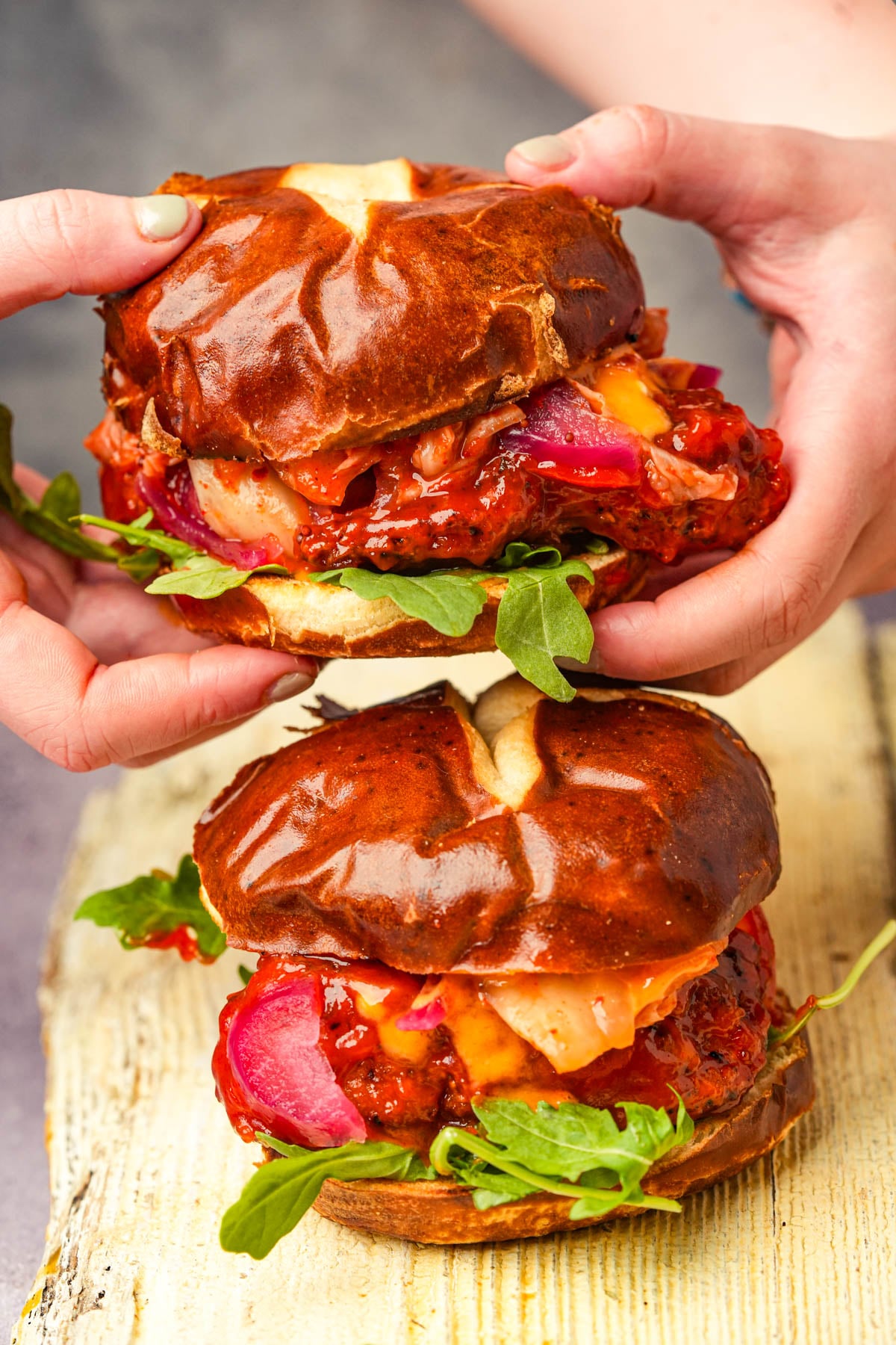 Two hands hold a large, overflowing gochujang seitan sandwich with glossy pretzel buns; a similar sandwich sits on a rustic wooden board below.