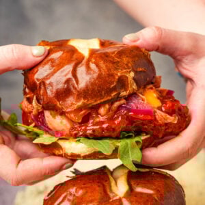 A person holds a gochujang seitan sandwich with a glossy pretzel bun.