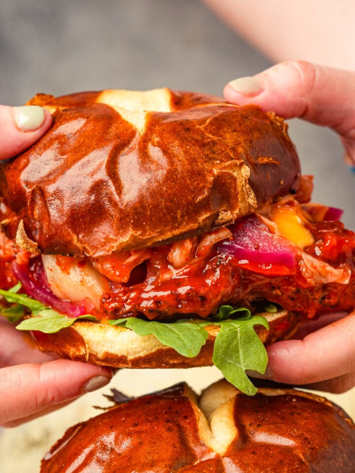 A person holds a gochujang seitan sandwich with a glossy pretzel bun.