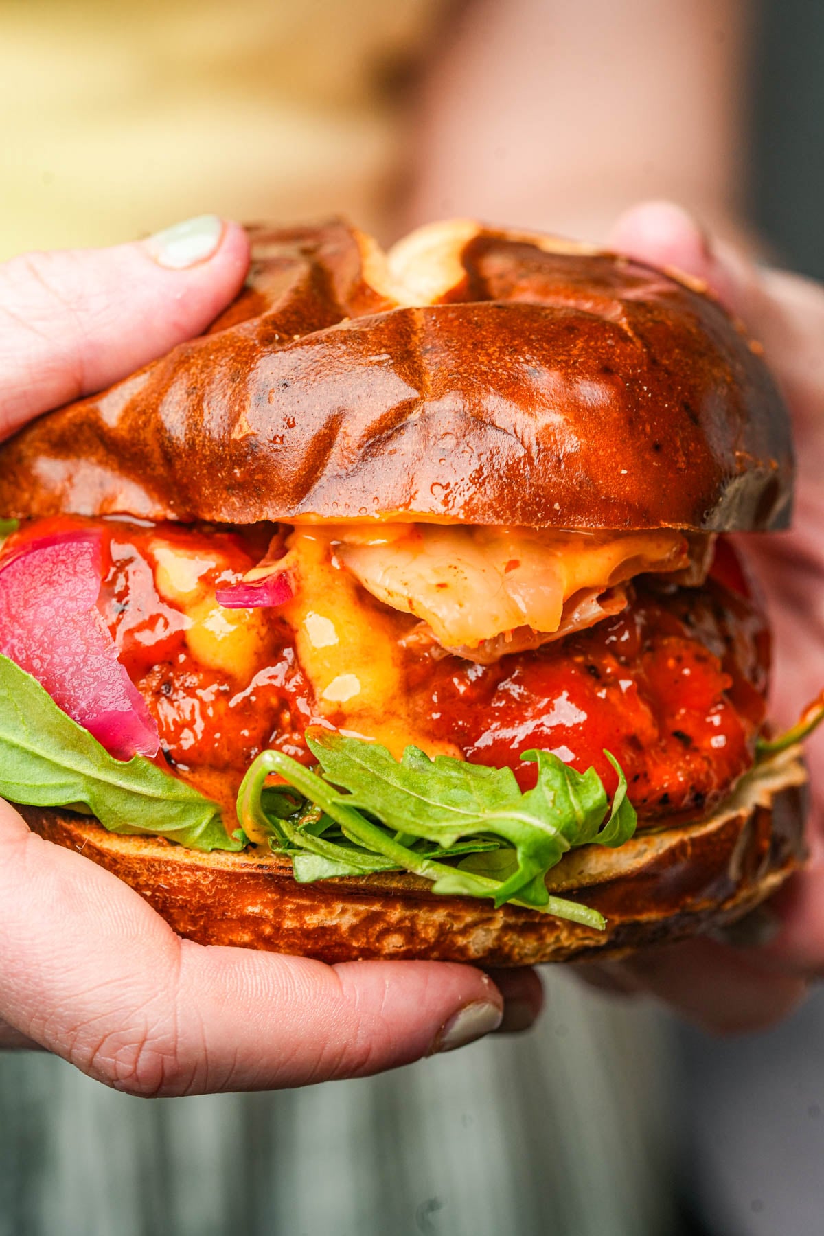 A person holds a large gochujang seitan sandwich filled with gochujang sauce-covered seitan chicken, sriracha mayo, pickled onions, kimchi, and fresh greens on a shiny pretzel bun.